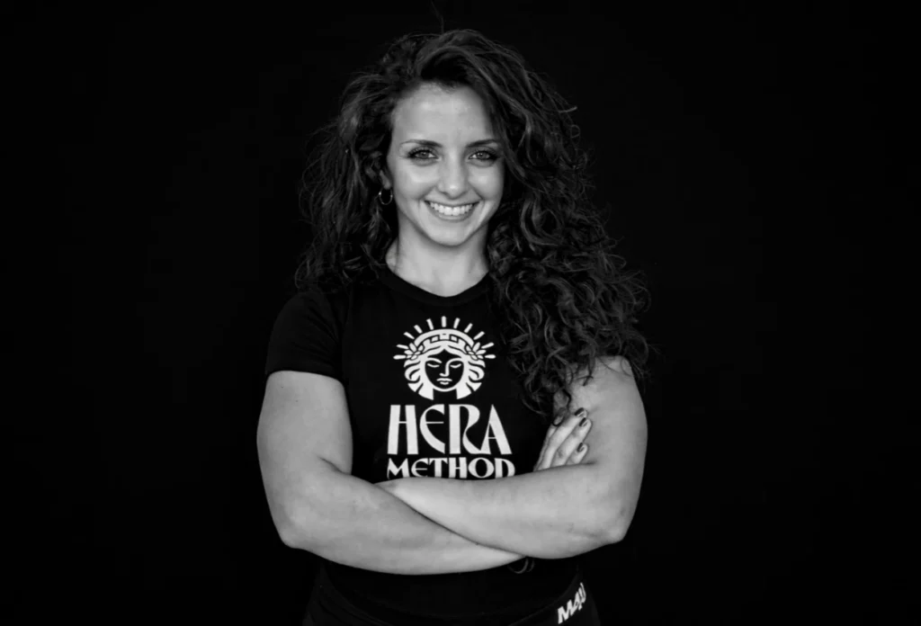 Woman with curly hair smiles, arms crossed, wearing a Hera Method shirt.
